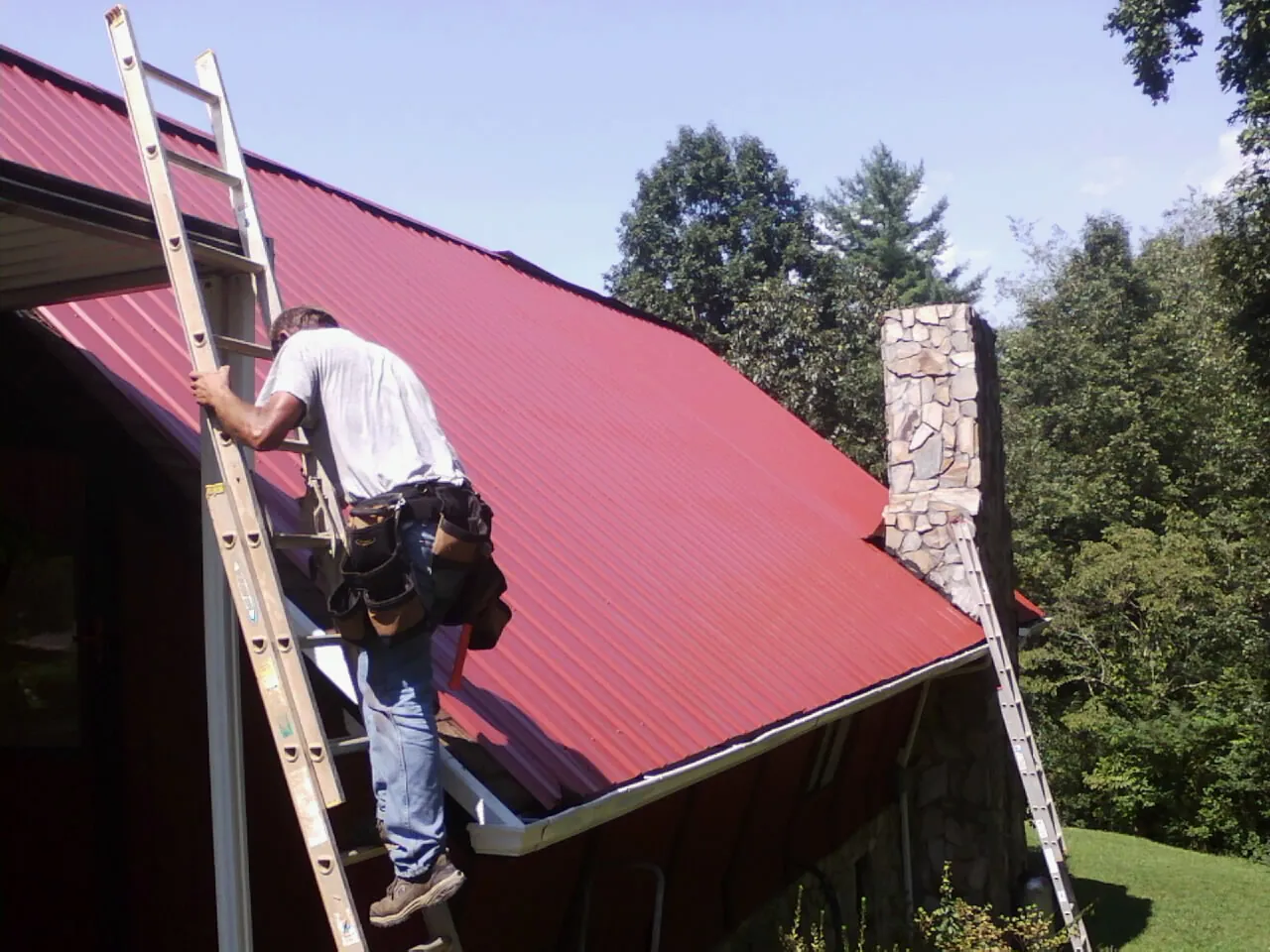 Roofer on a ladder beside a red metal roof and stone chimney — Soffit & Fascia Repair by Roofing Brothers in Adelanto